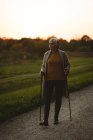 Seniorin im Morgengrauen mit Gehstöcken auf staubiger Straße unterwegs — Stockfoto