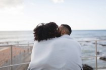 Vista trasera de pareja envuelta en un chal besándose cerca de la playa - foto de stock