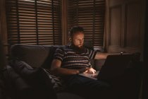 Homme dans la chambre noire assis sur le canapé en utilisant son ordinateur portable à la maison — Photo de stock