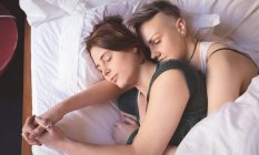 Lesbisches Paar hält Händchen, während es im Schlafzimmer zu Hause auf dem Bett schläft. — Stockfoto