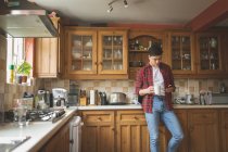 Homem usando telefone celular e segurando xícara de café na cozinha em casa . — Fotografia de Stock