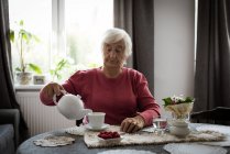 Seniorin gießt zu Hause Tee aus Teekanne in Tasse — Stockfoto