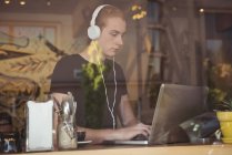 Hombre escuchando música en los auriculares mientras usa el ordenador portátil en la cafetería - foto de stock