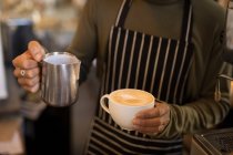 Barista tenant une tasse de café et une cruche de lait à la vapeur dans un café — Photo de stock