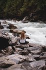Visão traseira da mulher sentada nas rochas perto de um rio fluindo — Fotografia de Stock