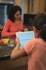Mädchen nutzt digitales Tablet, während Mutter zu Hause in der Küche Essen zubereitet — Stockfoto