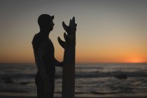 Surfista com prancha de surf em pé na praia ao pôr do sol — Fotografia de Stock