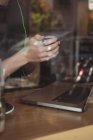 Средняя секция человека, пьющего кофе во время использования ноутбука в кафе — стоковое фото