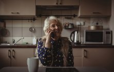 Улыбающаяся пожилая женщина разговаривает по телефону на кухне дома — стоковое фото