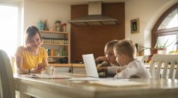 Mother and children using mobile phone and laptop in kitchen at home — Stock Photo