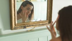 Mujer aplicando crema facial delante del espejo en casa . - foto de stock