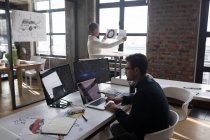 Бізнесмен використовує ноутбук під час колеги, дивлячись на ескіз в офісі . — стокове фото