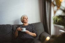 Старша жінка сидить на дивані, дивлячись далеко, тримаючи чашку кави вдома — стокове фото