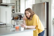 Женщина, использующая мобильный телефон, сохранив еду на кухне — стоковое фото
