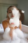 Женщина в маске для лица, выдувающая пену, принимая ванну в ванной . — стоковое фото