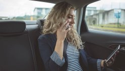 Улыбающаяся деловая женщина разговаривает по мобильному телефону при использовании цифрового планшета в автомобиле — стоковое фото