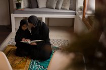 Madre musulmana ayudando a su hija a leer el Sagrado Corán en casa - foto de stock