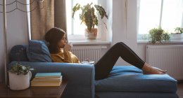 Молода жінка розслабляється на дивані з кавою в сонячний день вдома — стокове фото
