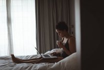 Молода жінка сидить на ліжку, використовуючи свій ноутбук, маючи каву в спальні вдома — стокове фото
