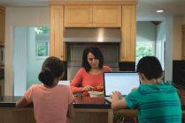 Madre preparando comida mientras los niños usan la tableta digital y el portátil en la cocina en casa - foto de stock