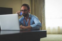 Empresário falando no celular enquanto usa laptop no quarto de hotel — Fotografia de Stock