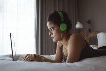 Donna che utilizza il computer portatile mentre ascolta musica sul letto in camera da letto — Foto stock