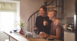 Happy lesbian couple using mobile phone and laptop while having coffee at home. — Stock Photo