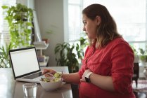 Беременная женщина завтракает дома, пользуясь ноутбуком — стоковое фото