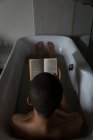 Junger Mann liest Buch, während er in Badewanne im Badezimmer liegt — Stockfoto