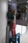 Junge auf Stuhl holt Essen aus Kühlschrank in Küche. — Stockfoto