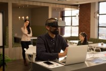 Мужчина-руководитель, использующий гарнитуру виртуальной реальности во время работы над ноутбуком в кафетерии — стоковое фото