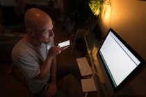 Человек, работающий на персональном компьютере во время использования мобильного телефона дома . — стоковое фото