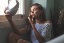 Woman applying lipstick in front of mirror near window at home. — Stock Photo