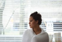 Nachdenkliche Frau in Schal gehüllt, die zu Hause aus dem Fenster schaut — Stockfoto