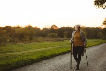 Donna anziana che cammina su una strada polverosa con bastoni all'alba — Foto stock