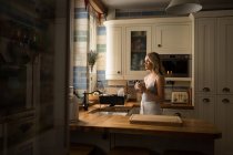 Vista lateral de la joven mujer de pie en la cocina sosteniendo taza - foto de stock