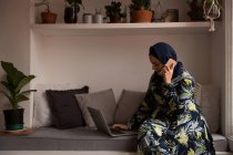 Mulher muçulmana falando ao telefone enquanto usa laptop em casa — Fotografia de Stock
