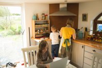Mother and son preparing food while daughter using laptop in background in kitchen at home — Stock Photo