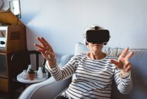 Femme âgée assise sur un canapé portant un casque de réalité virtuelle à la maison — Photo de stock