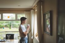 Young man drinking coffee while looking through window at home. — Stock Photo