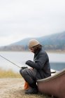 Человек, сидящий на лодке со своим рыболовецким снаряжением возле реки — стоковое фото