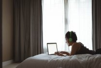 Woman using laptop while listening to music on bed in bedroom — Stock Photo
