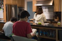 Crianças lendo livro enquanto a mãe se prepara na cozinha em casa — Fotografia de Stock