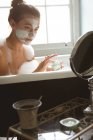 Frau trägt Gesichtsmaske vor Spiegel auf, während sie zu Hause baden geht. — Stockfoto