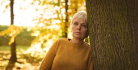 Nachdenkliche Seniorin lehnt an einem sonnigen Tag im Park an einem Baumstamm — Stockfoto