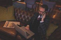 Бизнесмен берет на себя мобильный телефон во время использования ноутбука в баре — стоковое фото