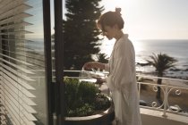 Junge lächelnde Frau, die an einem sonnigen Tag auf dem Balkon steht und die Pflanzen zu Hause gießt — Stockfoto