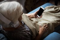 Donna anziana che ascolta musica con le cuffie mentre utilizza il telefono cellulare in soggiorno — Foto stock