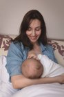 Улыбающаяся молодая мать, сидящая на кровати и кормящая грудью ребенка дома — стоковое фото