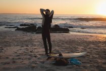 Vista traseira do surfista em pé na praia durante o anoitecer — Fotografia de Stock